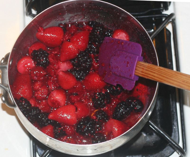 A pot with strawberries, blackberries, raspberries and sugar on the stove top. A spatula is being used to stir the berries.