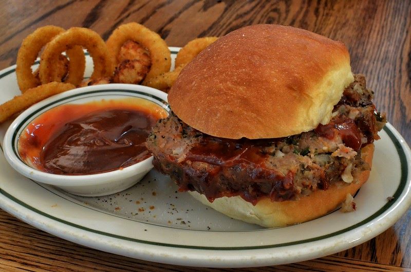 A plate with a meatloaf sandwich in a bun, onion rings, and a small dish with ketchup.