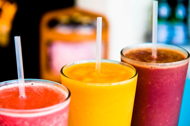 Three smoothies in glasses with straws. The first one is pink, the second is yellow, and the third is purple.