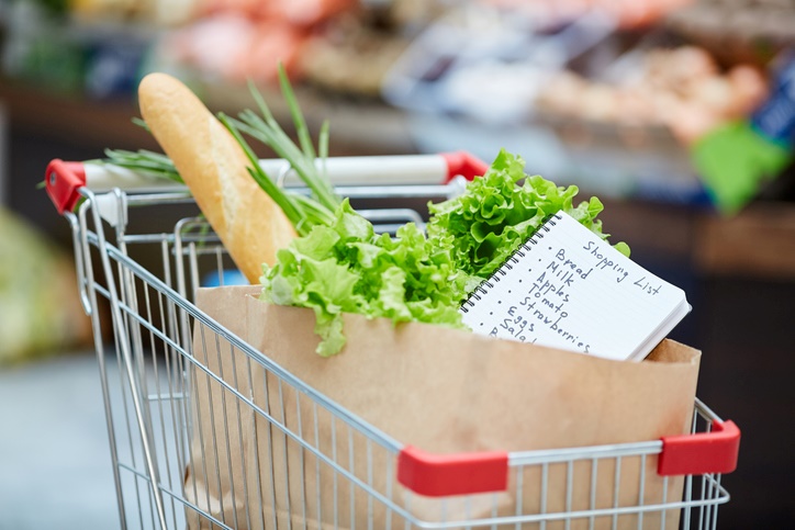 A shopping cart filled with groceries, with a shopping list sitting on top