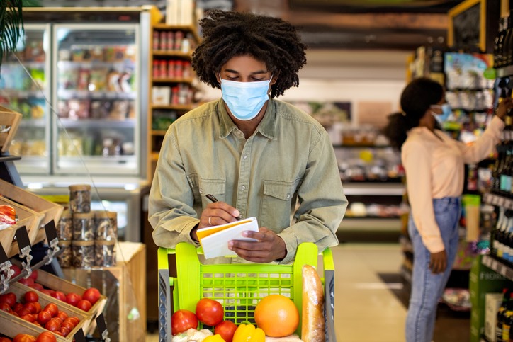 A person with a cart at the grocery store, looking down at a shopping list